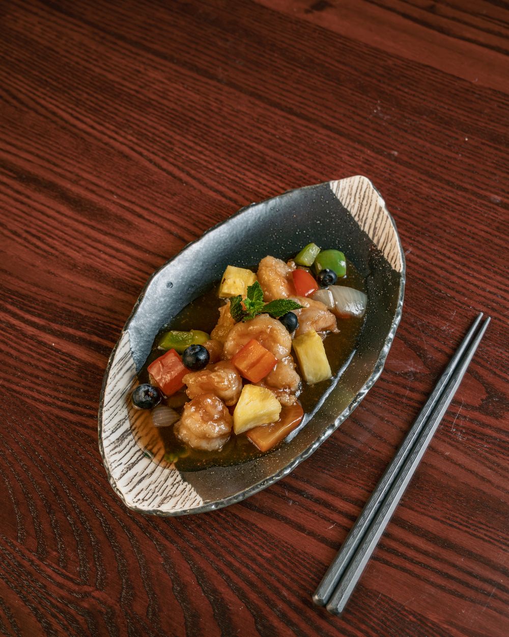 Sweet and sour king prawns with pineapple and peppers in a dark ceramic dish.