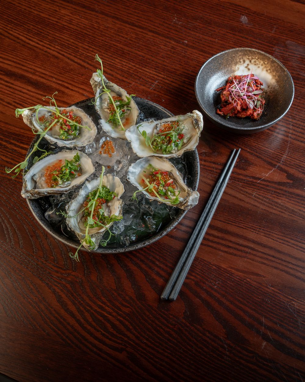 Six kimchi oysters on crushed ice, with a small dish of kimchi beside them.