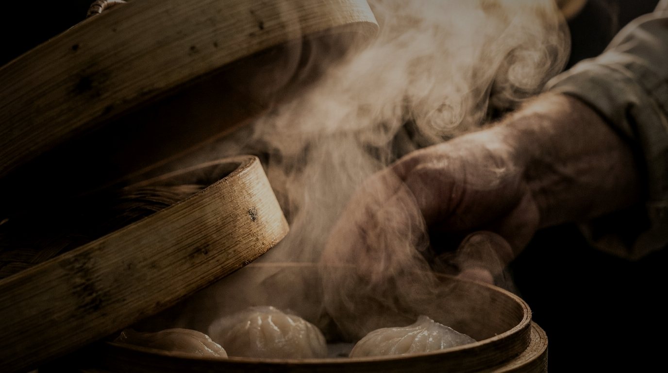 A bamboo steamer lifted open, steam rising across a chef's hand and half hiding the dumplings beneath.